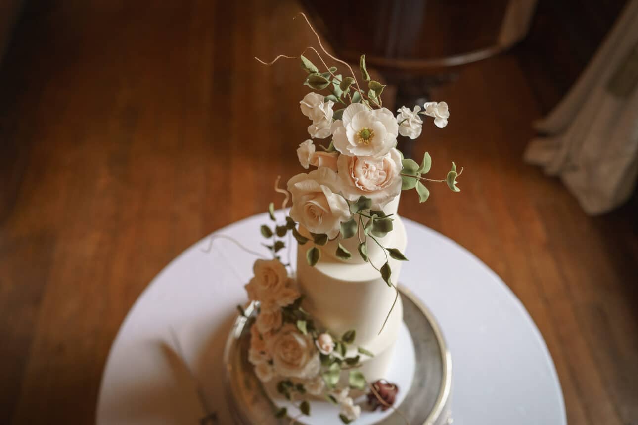 a two tiered white cake decorated with white and pale peach flowers and greenery, placed on a silver tray at a sandon hall wedding, with a cake knife beside it on a round table.