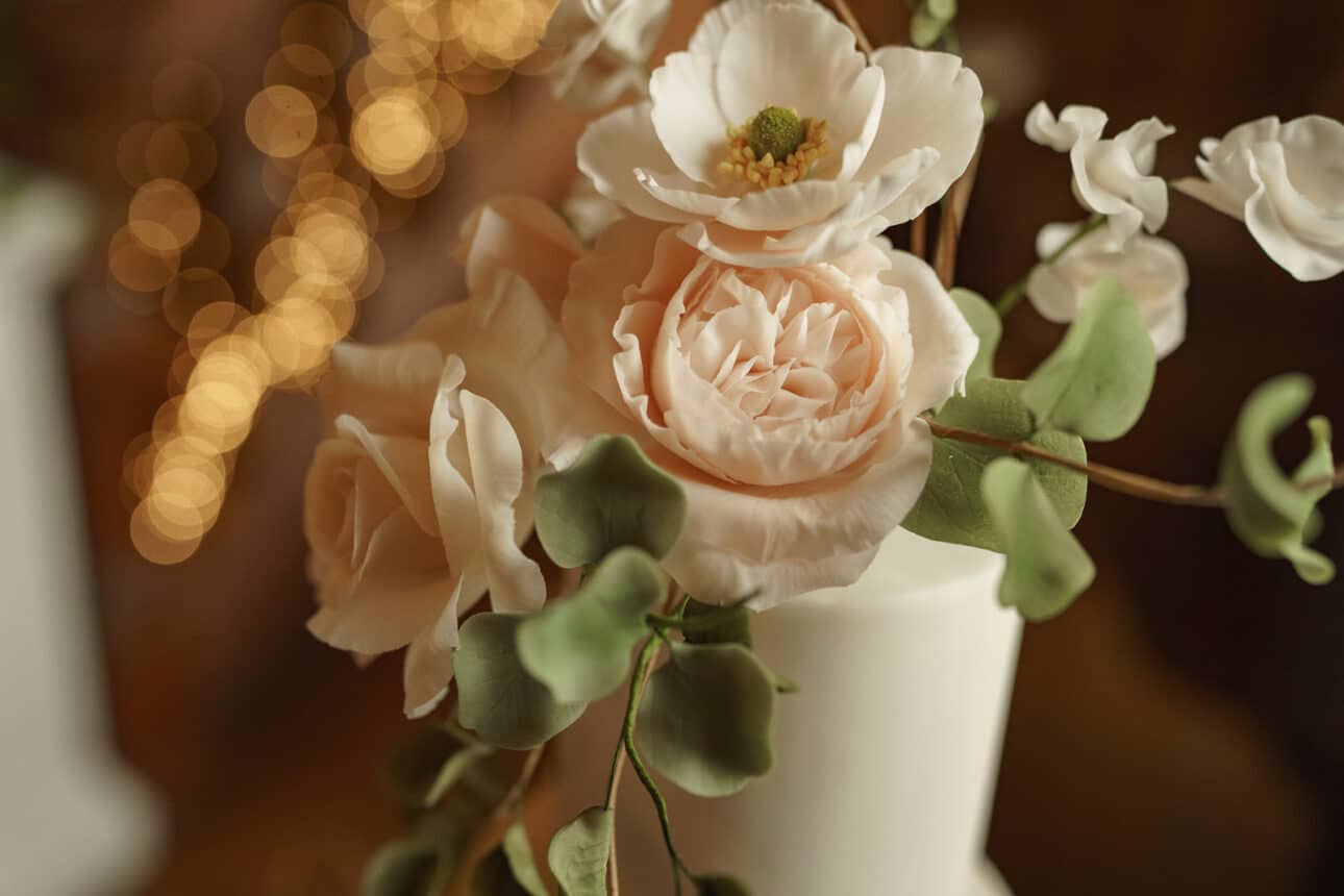 close up of a white cake decorated with realistic sugar flowers and green leaves, glowing under warm, blurred lights—the perfect centerpiece for an elegant sandon hall wedding.