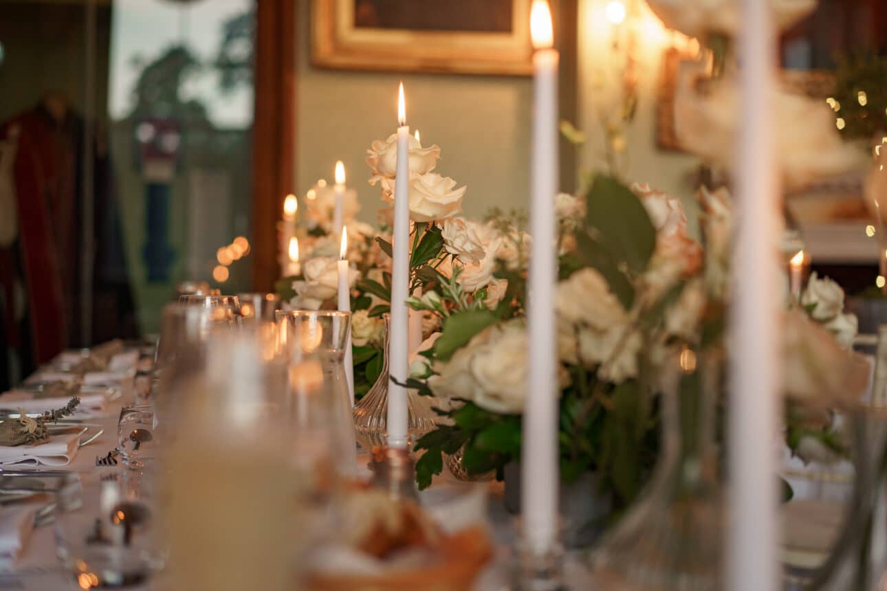 a formal table setting at sandon hall wedding features white roses, tall lit candles, glassware, and plates elegantly arranged for a meal in a warmly lit room.