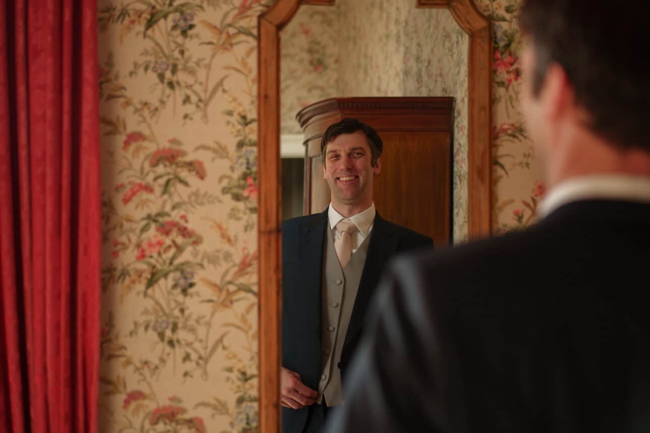 a man in formal attire stands in front of a mirror, smiling at his reflection in a room with floral wallpaper and red curtains at sandon hall.