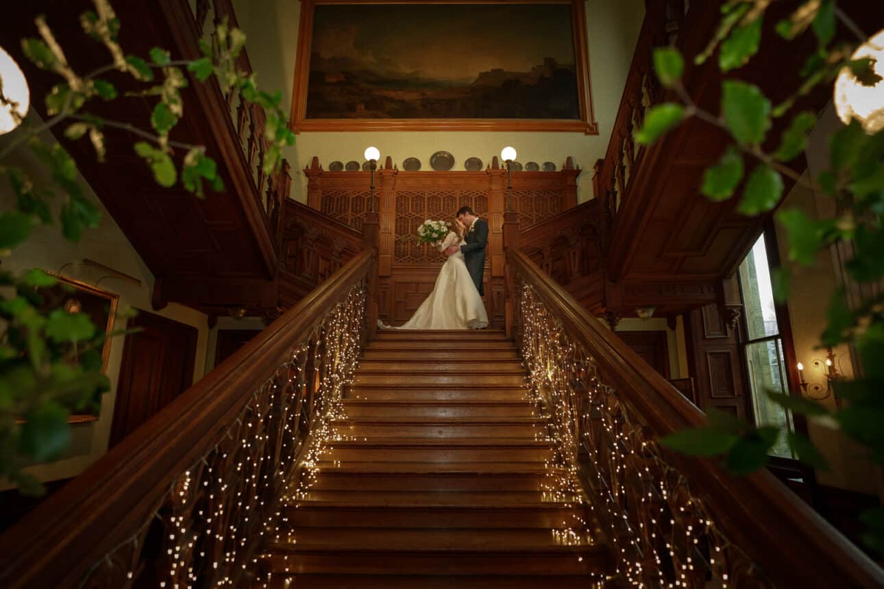 a bride and groom stand at the top of a grand wooden staircase at sandon hall, decorated with string lights and surrounded by ornate architecture and greenery, capturing the magic of a sandon hall wedding.