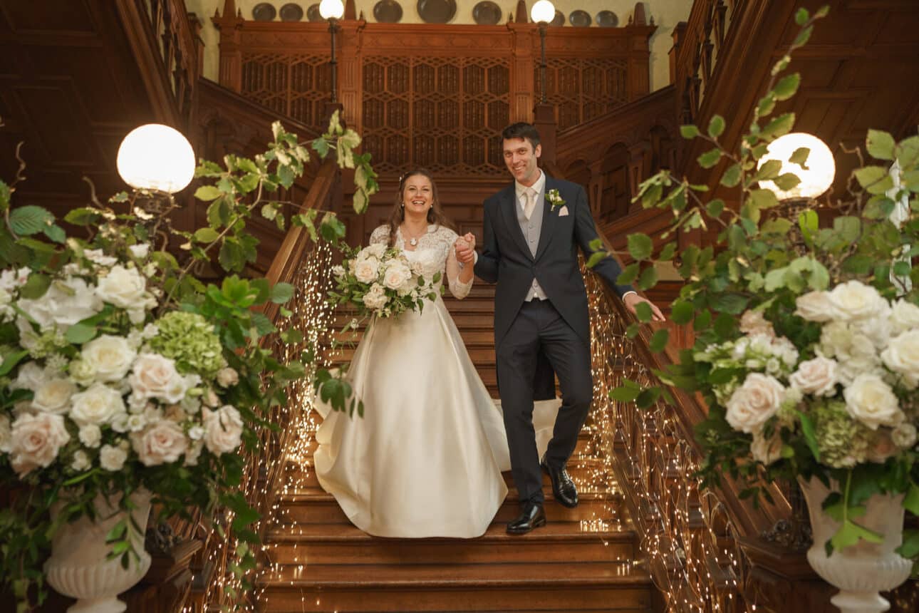 a bride and groom walk down a grand staircase decorated with flowers and lights inside the ornate wooden venue of sandon hall, creating a magical sandon hall wedding atmosphere.