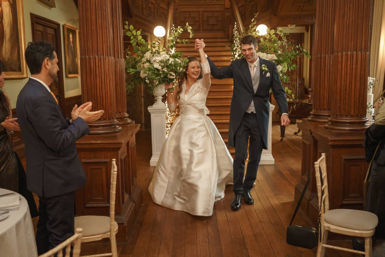 a bride and groom enter the wooden hall at sandon hall holding hands, smiling, while guests applaud.