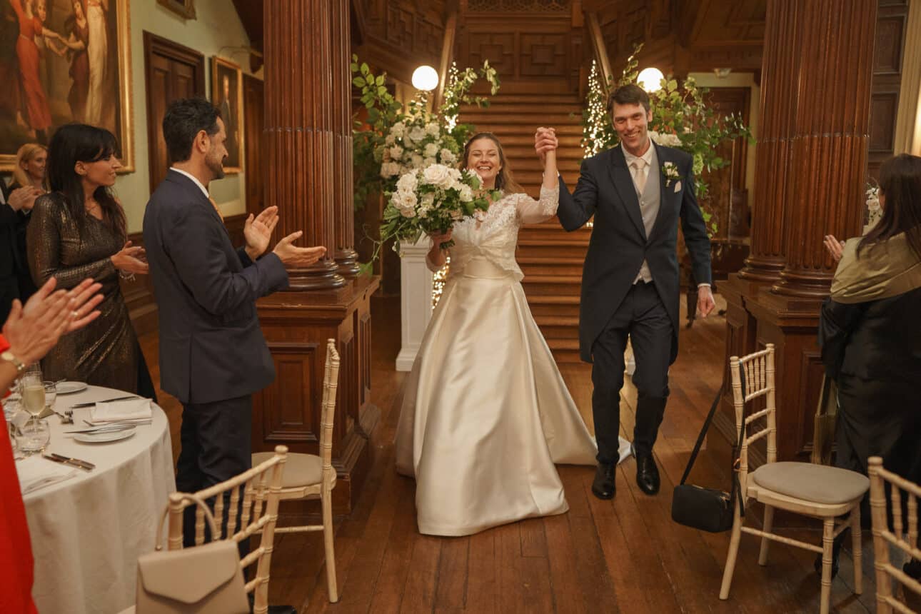 a bride and groom enter the wood paneled sandon hall holding hands and smiling as guests stand and clap. the bride holds a large bouquet of white flowers at their sandon hall wedding.