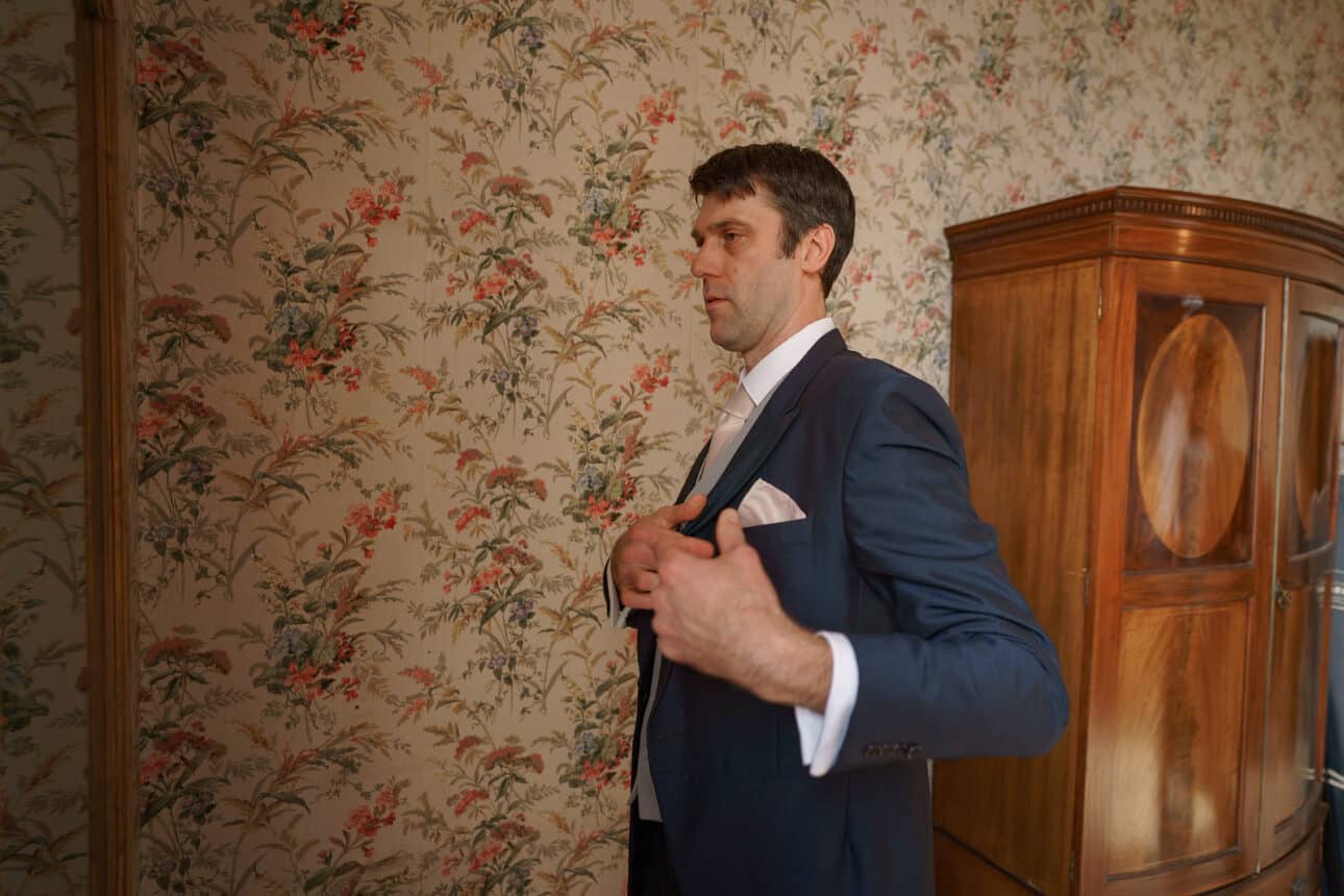 a man in a blue suit adjusts his jacket while standing in a room with floral wallpaper and a wooden wardrobe at a sandon hall wedding.