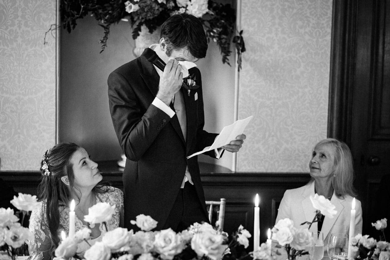 a man in a suit wipes his eyes while reading a paper, with two women beside him at a decorated table during a formal sandon hall wedding.