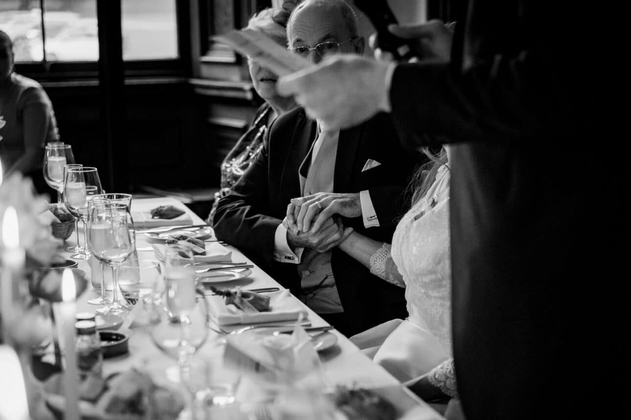 a black and white photo captures seated wedding guests holding hands at a decorated table during a sandon hall wedding, while another person stands nearby holding a piece of paper, possibly giving a speech.