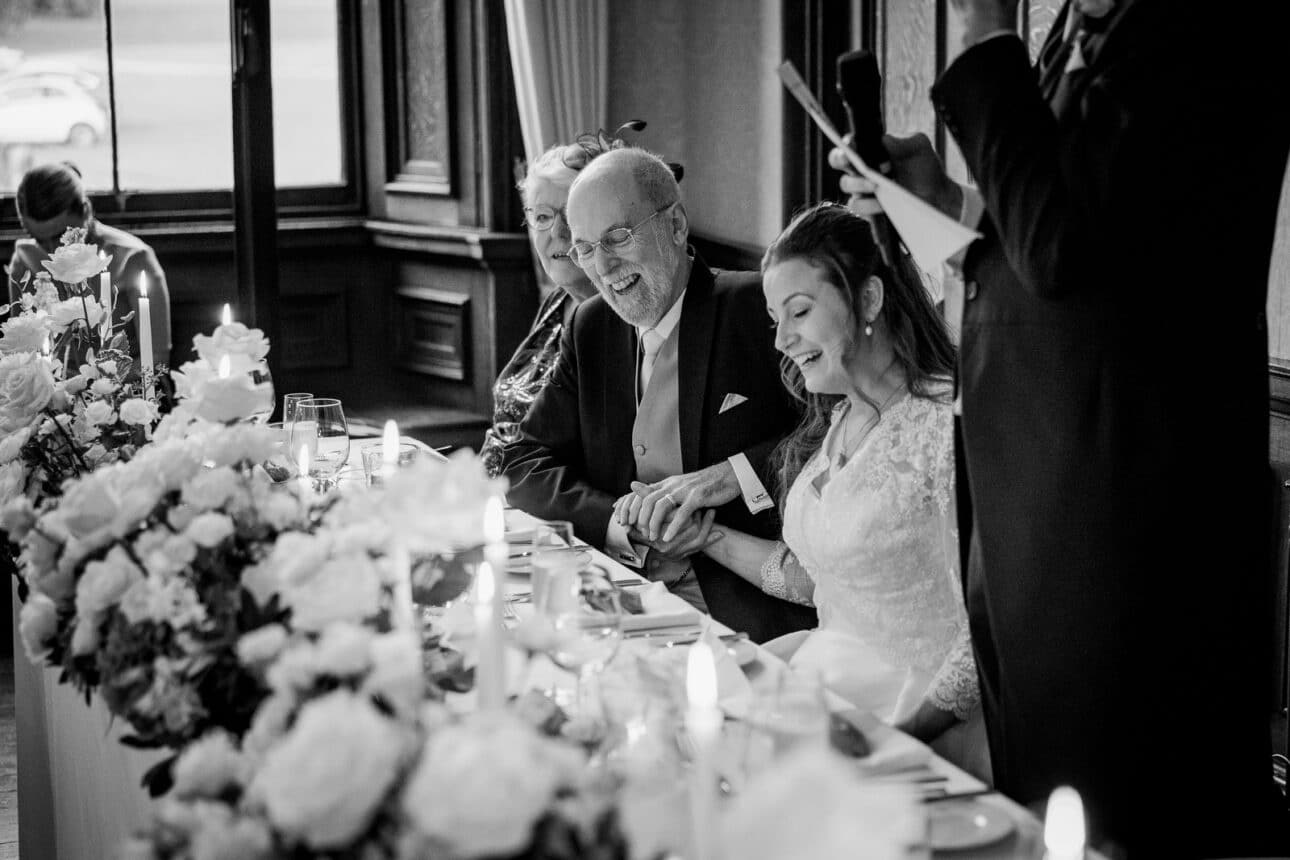 a bride and two older guests sit at a decorated sandon hall wedding table, smiling and holding hands, while someone stands nearby reading from a piece of paper.