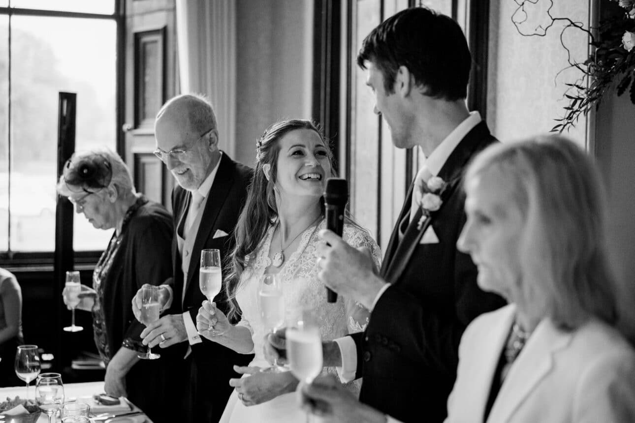a bride and groom stand with family at a sandon hall wedding reception table, holding glasses. the groom speaks into a microphone while the bride smiles at him.