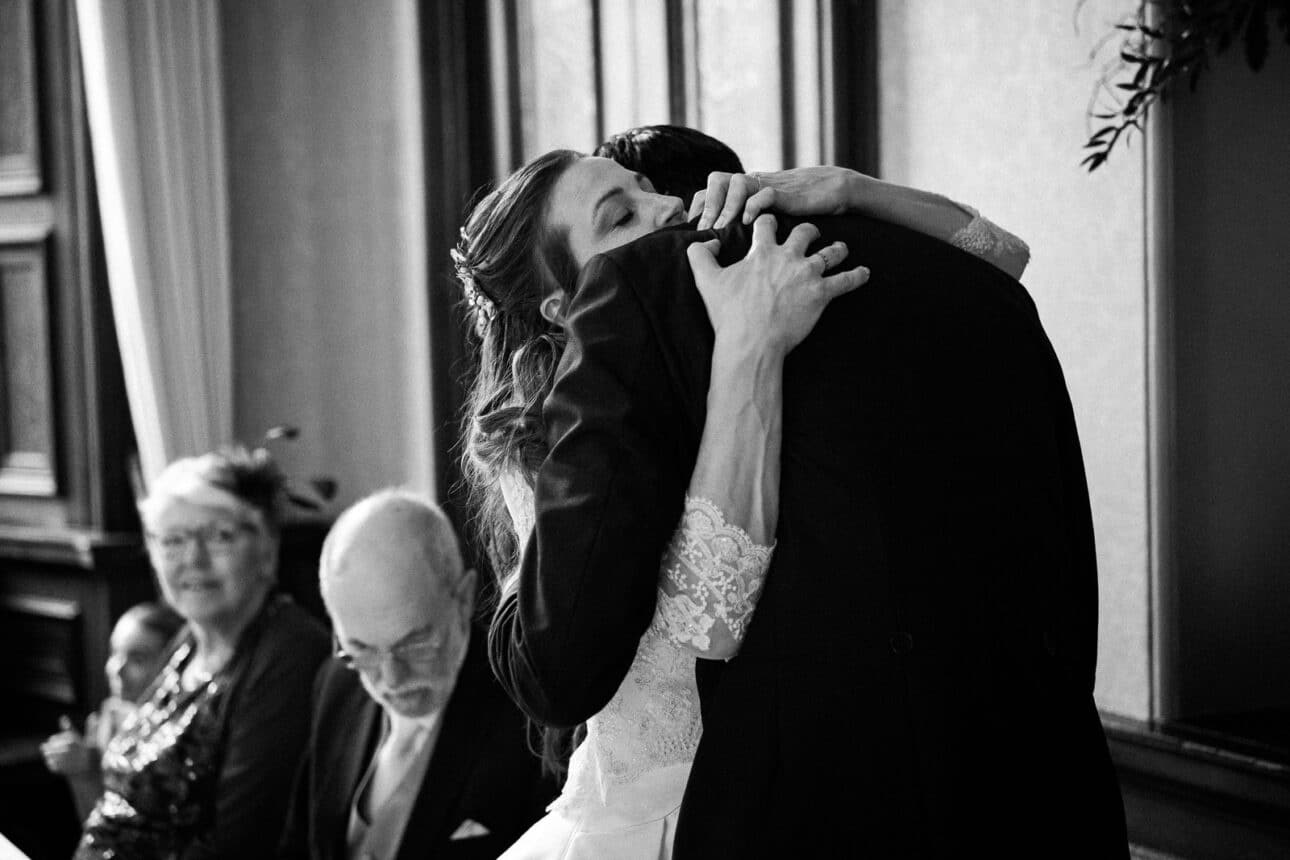 a bride and groom embrace tightly indoors at their sandon hall wedding, while two seated guests look on and another writes at a table.