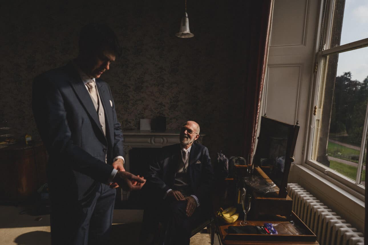 two men in suits, one standing and the other seated, are in a dimly lit room near a large window with outdoor scenery visible, capturing a quiet moment at a sandon hall wedding.
