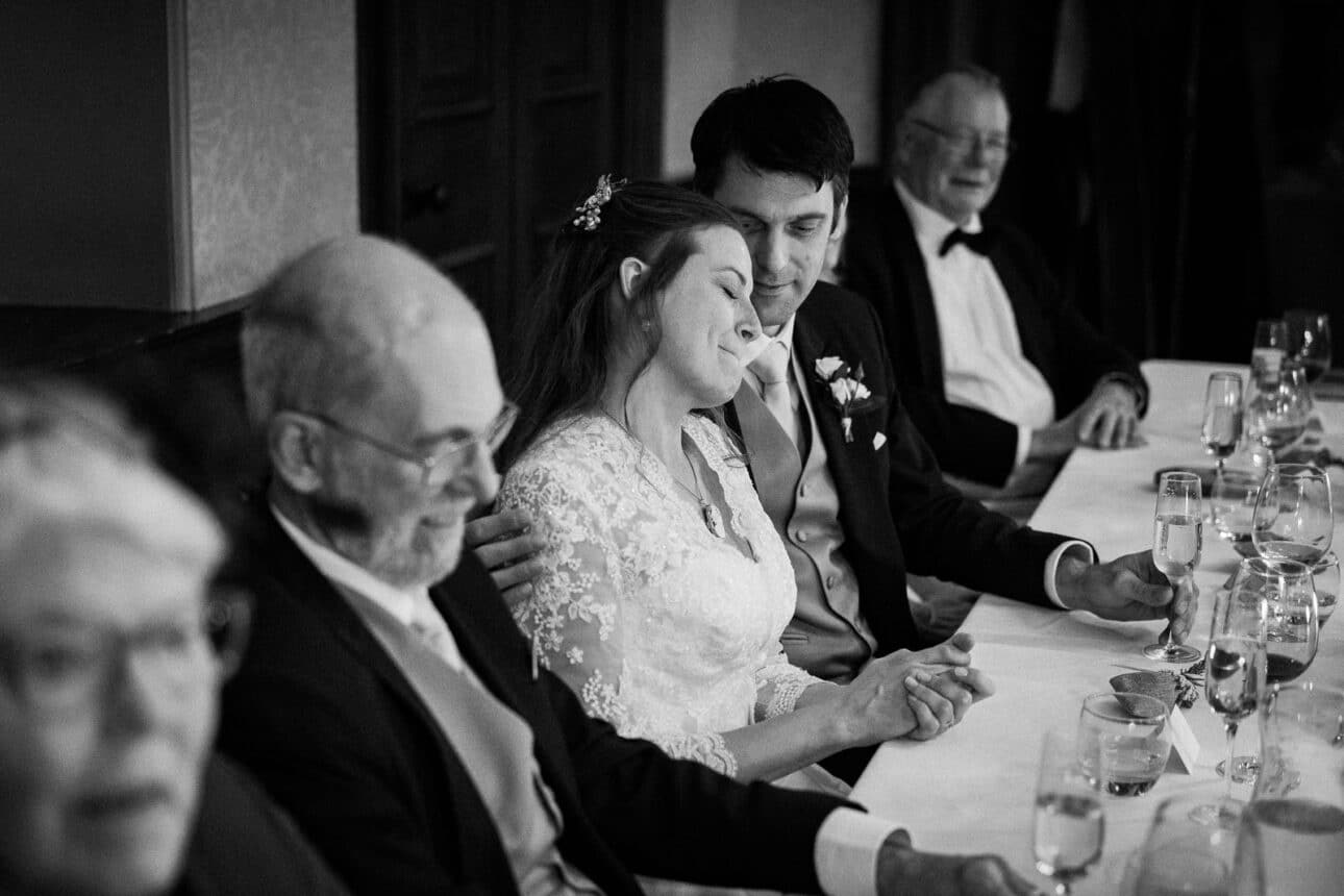 a bride and groom sit closely together at a banquet table, holding hands and smiling, surrounded by other formally dressed guests at their elegant sandon hall wedding.