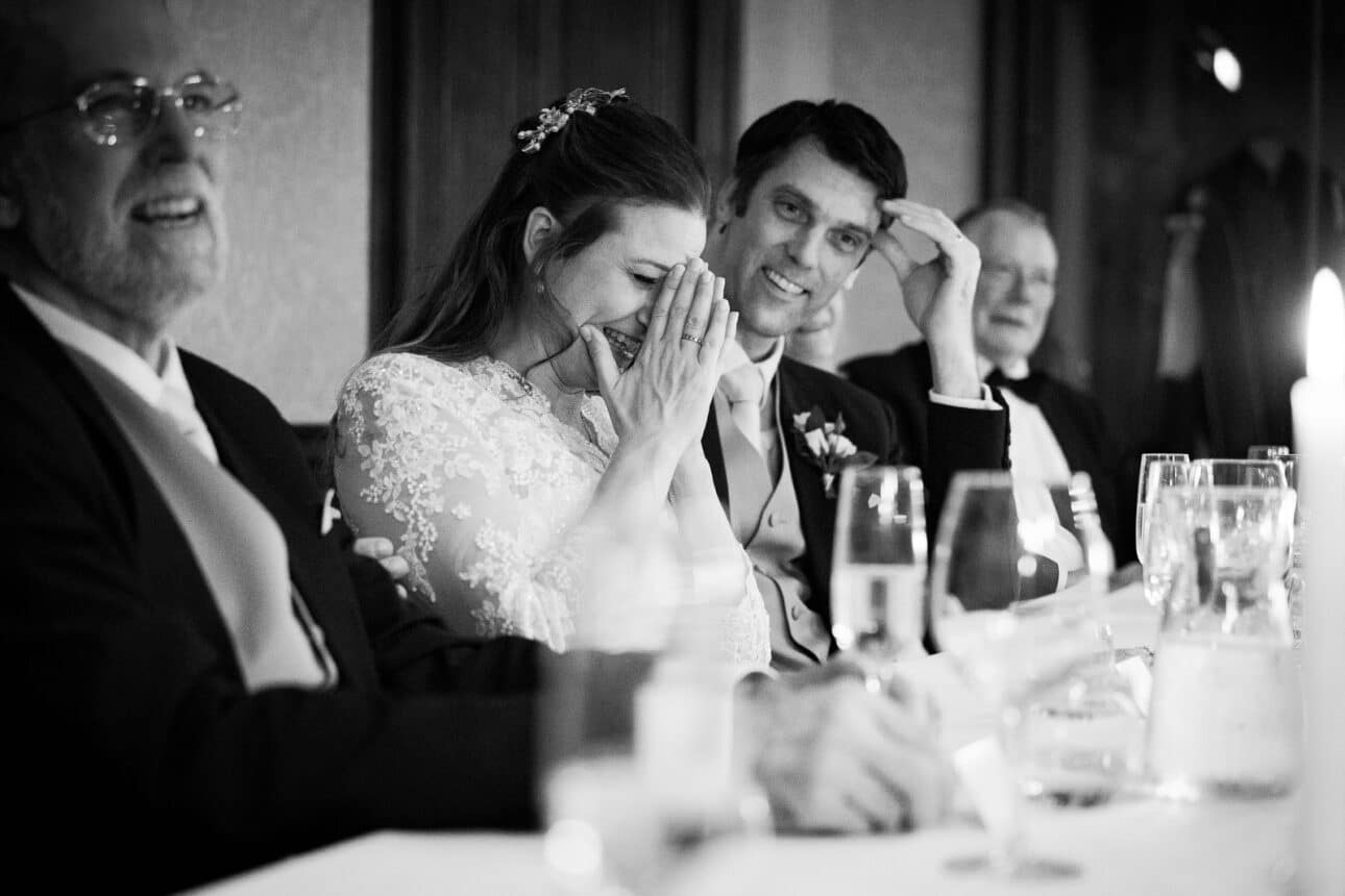 a bride and groom sit at a table at their sandon hall wedding, the bride laughing with her hands covering her face, surrounded by two male guests in formal attire at the reception.