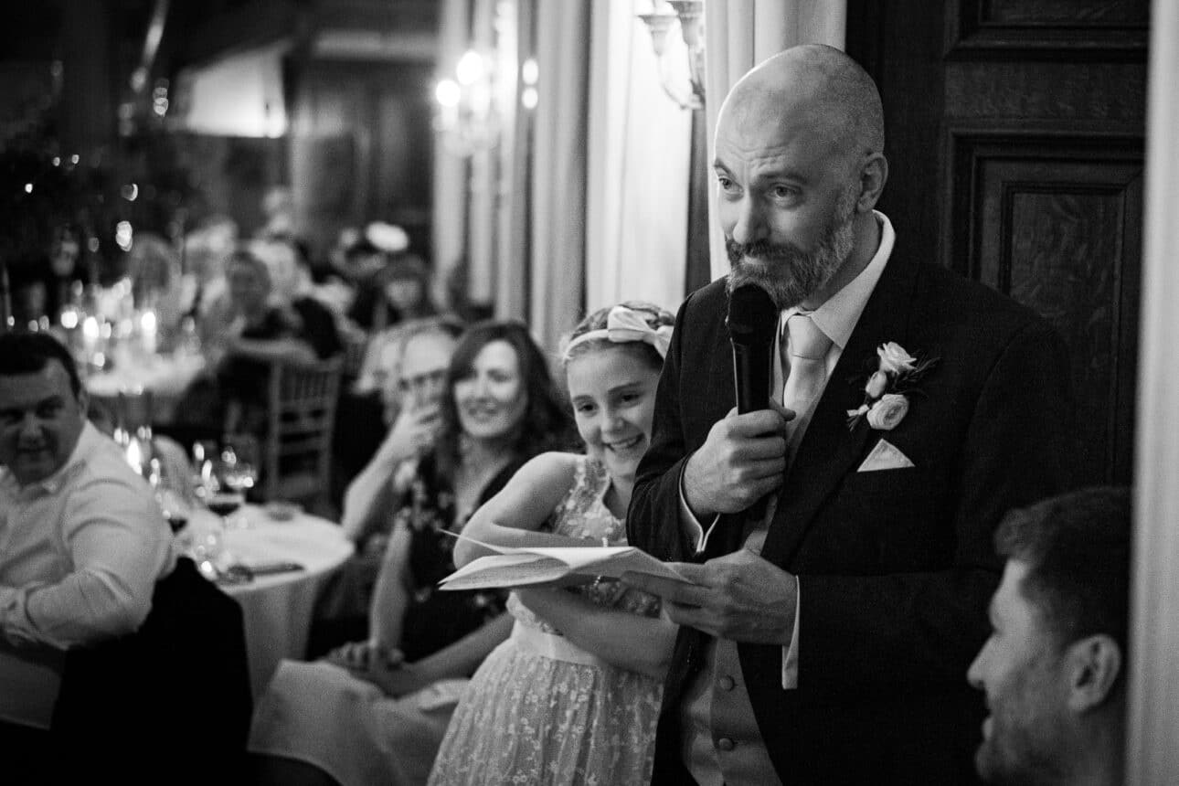 at a sandon hall wedding, a man in a suit speaks into a microphone and holds papers, while a young girl stands behind him smiling; guests seated at tables watch.