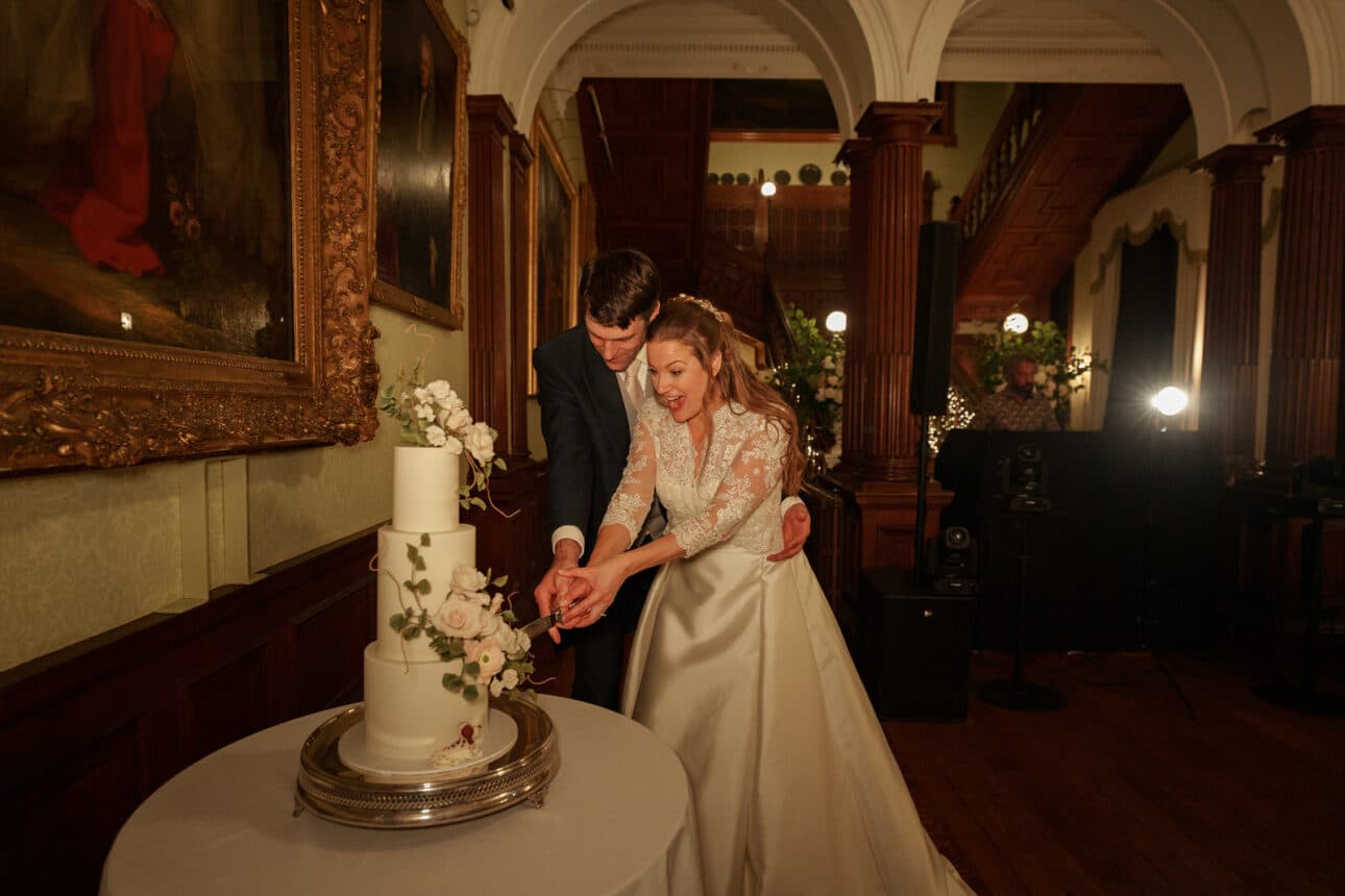 a bride and groom in formal attire cut a white, tiered wedding cake together in an ornate, warmly lit room at their elegant sandon hall wedding.