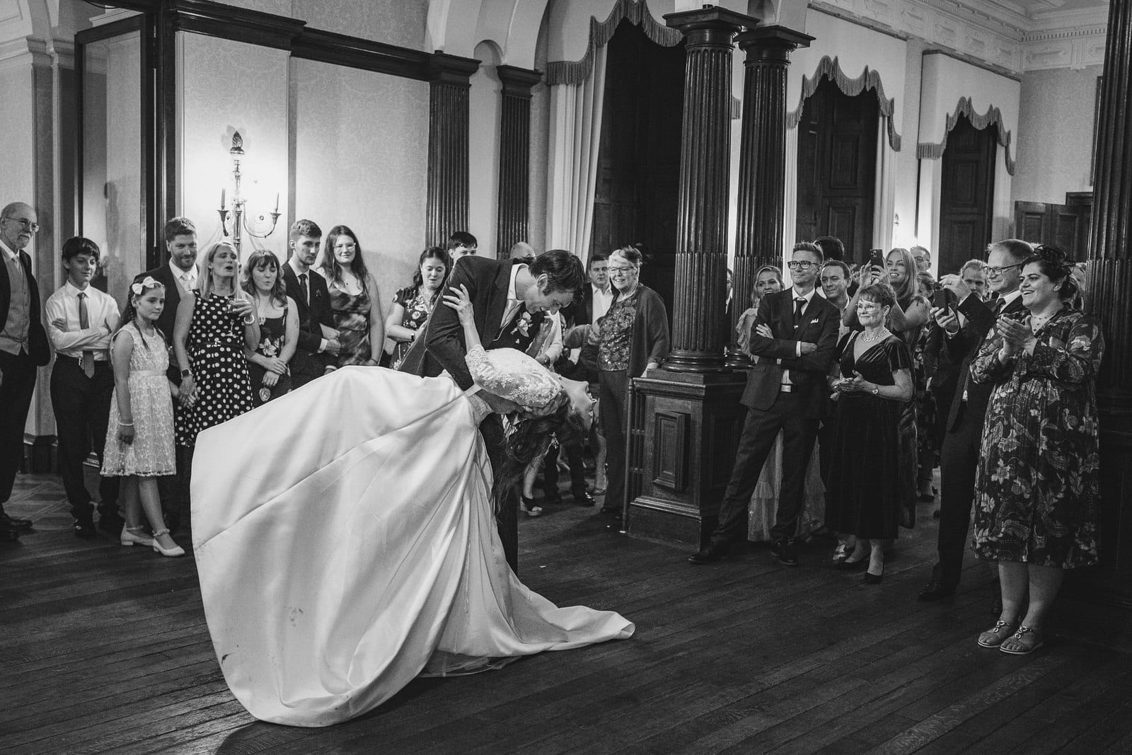 a bride and groom share a kiss on the dance floor while wedding guests watch in an elegant, formal room at sandon hall.