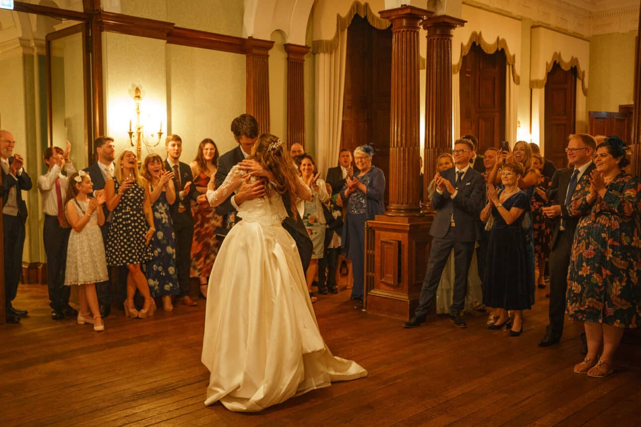 two women in wedding dresses embrace and dance in the center of a sandon hall wedding, surrounded by guests who are clapping and watching.