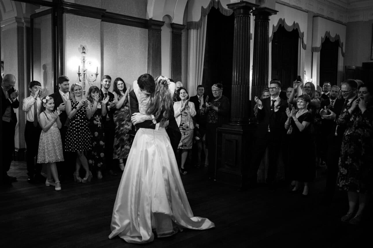a bride and groom share a kiss on the dance floor while guests stand around them, clapping and smiling in a formal, elegant sandon hall wedding setting.