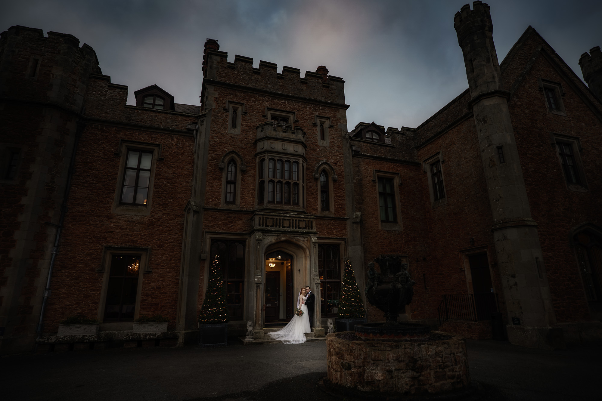 A bride and groom stand at the entrance of a large, historic stone building with turrets and dim evening lighting, beautifully captured by a talented Shropshire wedding photographer.