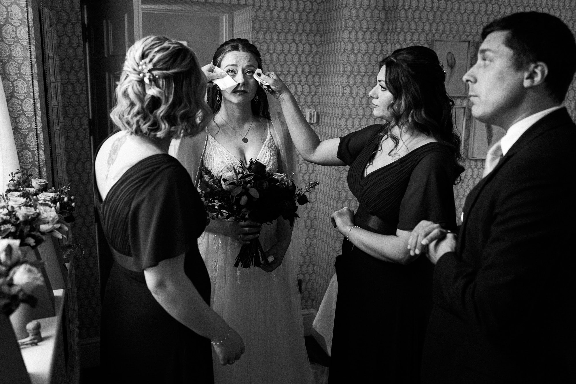 a bride in a wedding dress has her makeup touched up by two bridesmaids, while holding a bouquet, as a man in a suit stands nearby in a decorated room—captured beautifully by pbartworks photography, leading shropshire wedding photographer.