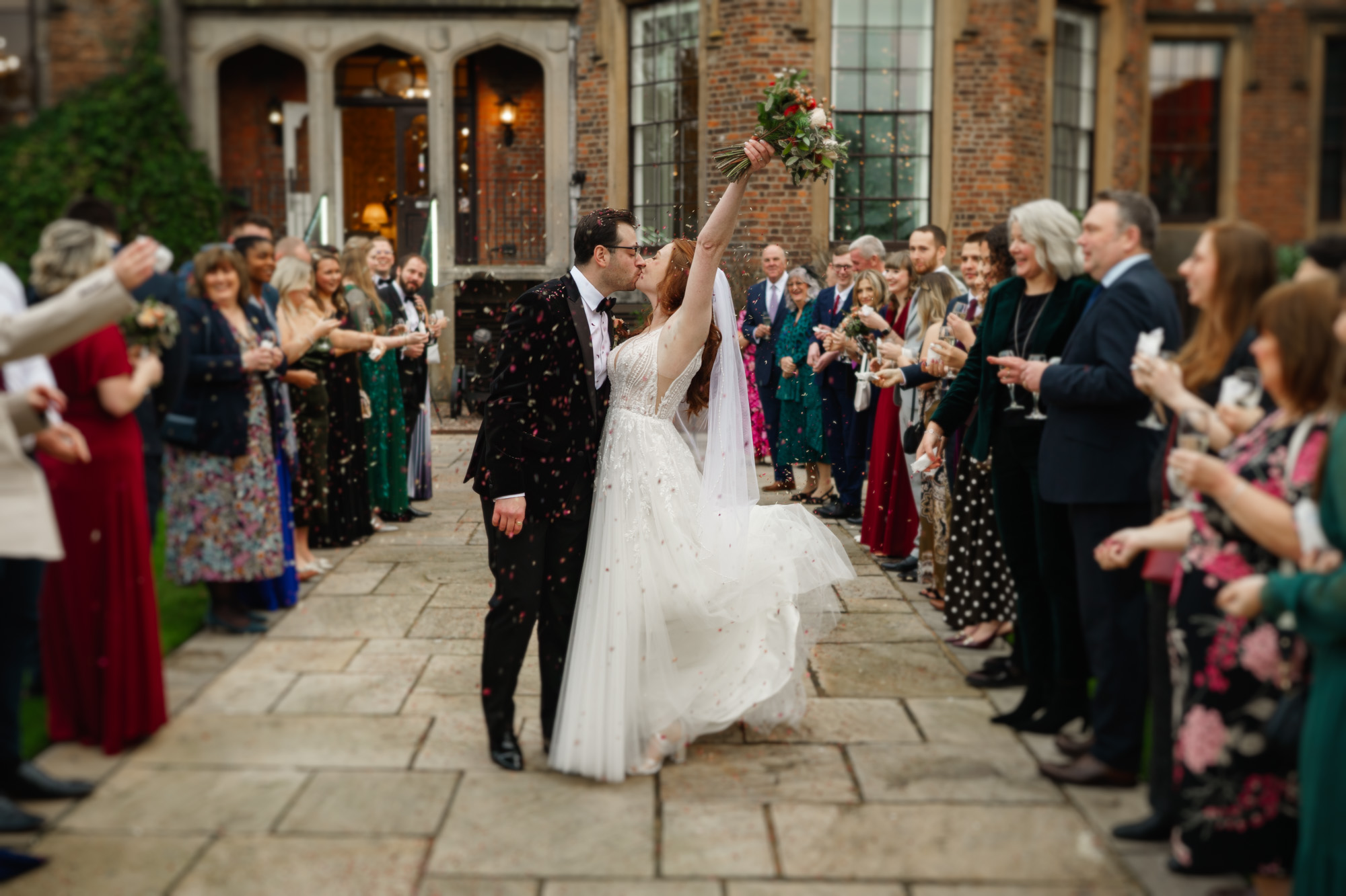 a bride and groom kiss outdoors as guests shower them with confetti. the bride raises her bouquet in celebration—a perfect moment captured by a wedding photographer in shropshire.
