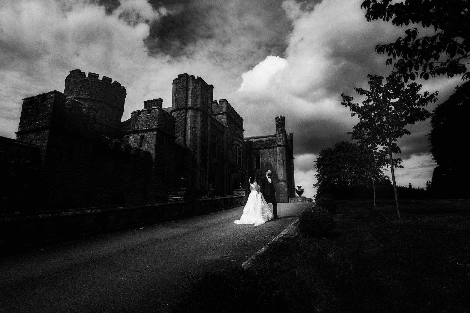 a bride and groom stand on a drive in front of a large, old stone castle under a dramatic cloudy sky—a perfect scene for a shropshire wedding photographer to capture unforgettable memories.
