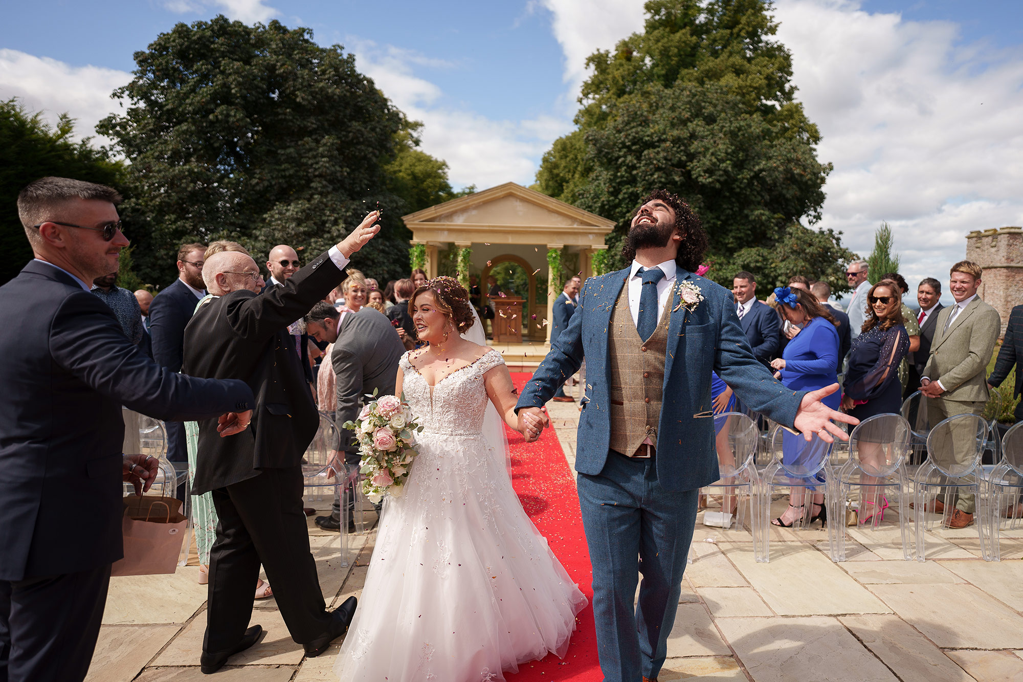 a bride and groom walk down a red carpet outdoors, smiling and celebrating as guests on both sides applaud and throw confetti—beautifully captured by a shropshire wedding photographer.