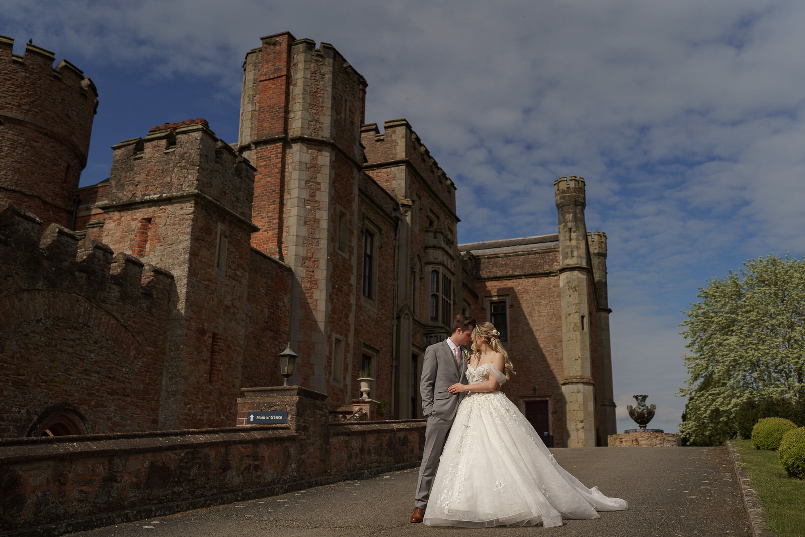 a bride and groom stand close together on a paved path outside a large stone castle on a partly cloudy day, beautifully captured by shropshire wedding photographer pbartworks photography.