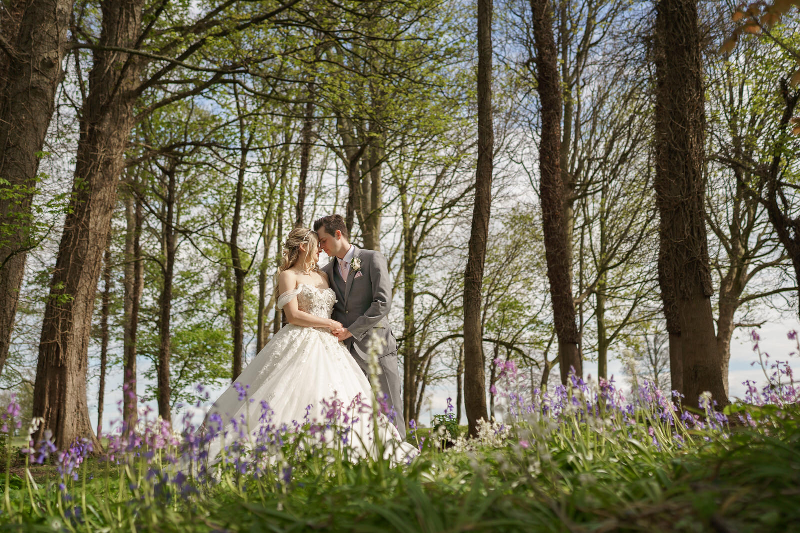a bride in a white ballgown and groom in a grey suit stand together among bluebells in a woodland setting, touching foreheads—captured beautifully by pbartworks photography, renowned wedding photographers in shropshire.