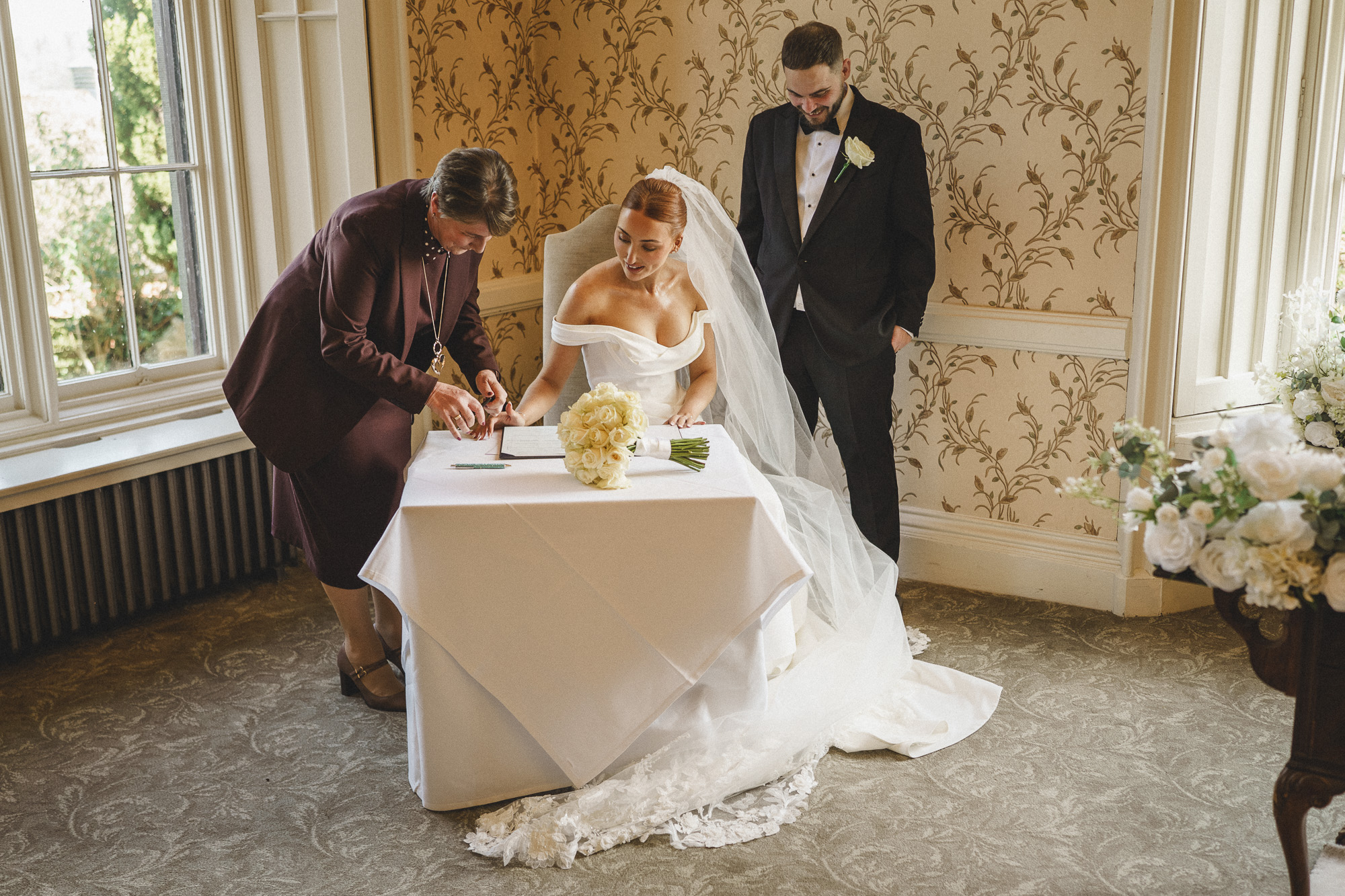 a bride signs a document at a table, with a bouquet nearby, while an officiant points and the groom stands beside them—captured beautifully by pbartworks photography, trusted wedding photographers in shropshire.