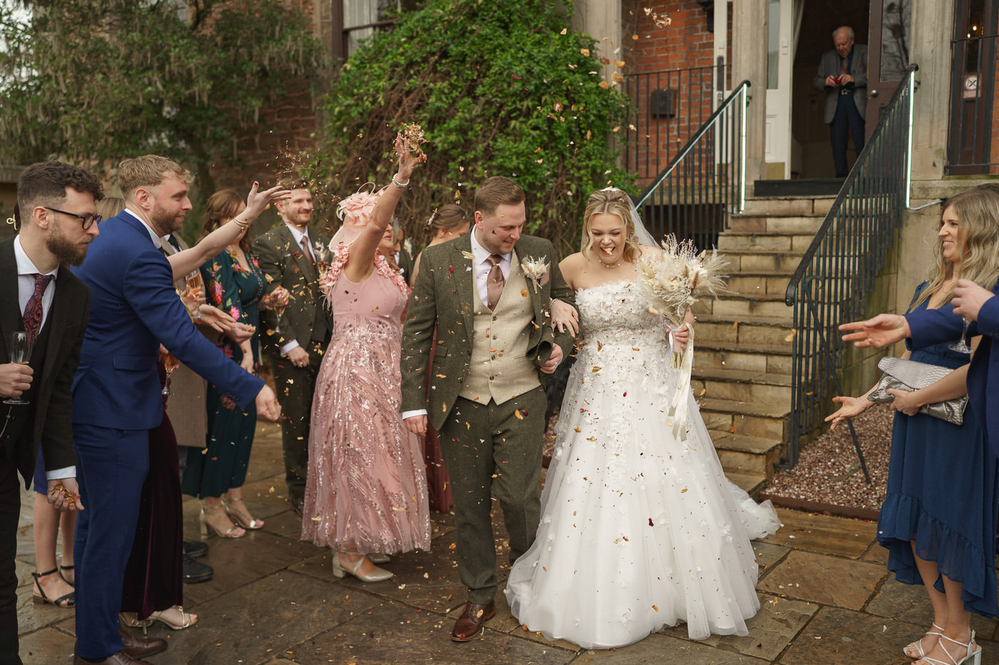 a bride and groom walk outdoors in wedding attire while guests throw confetti at them and smile, beautifully captured by a shropshire wedding photographer.