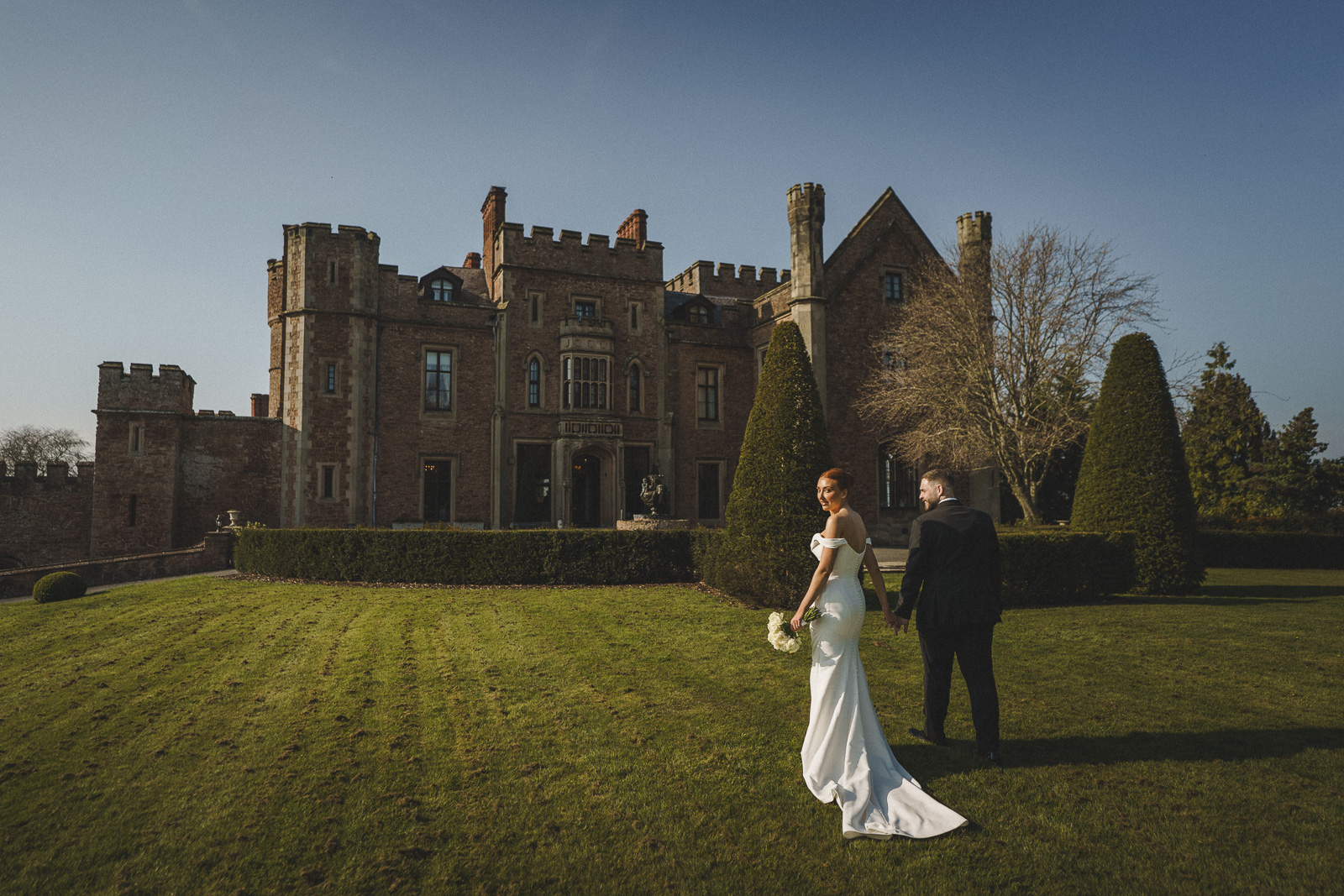 a bride and groom stand on the lawn before a grand stone castle on a clear day, the bride holding a bouquet—captured by shropshire wedding photographer pbartworks photography.