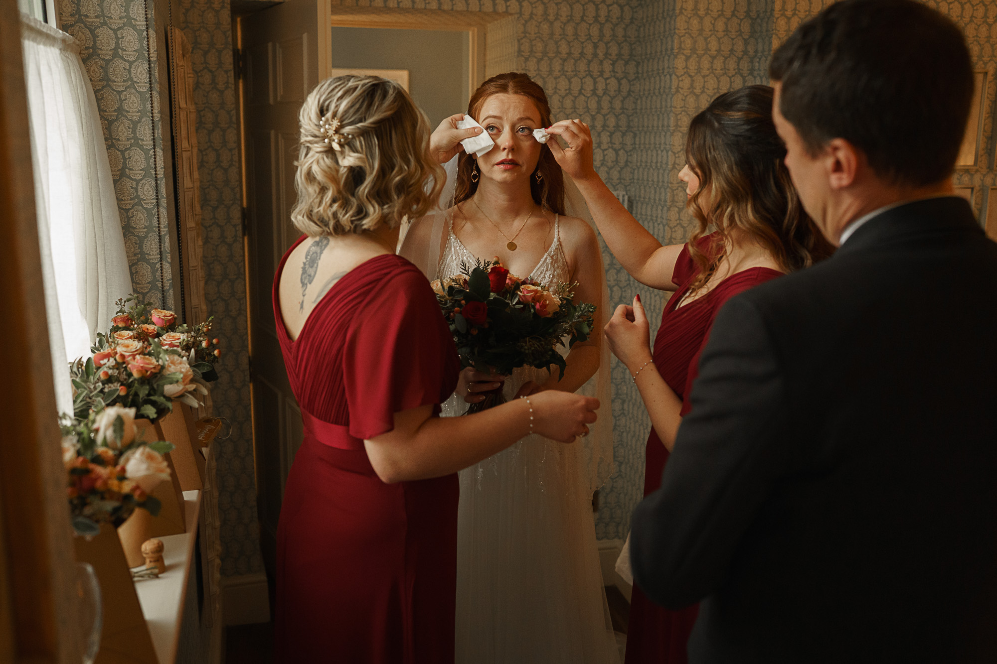 a bride holding a bouquet stands still while two women in red dresses help her blot tears; a man in a suit stands nearby in a softly lit room, perfectly captured by pbartworks photography, a trusted shropshire wedding photographer.