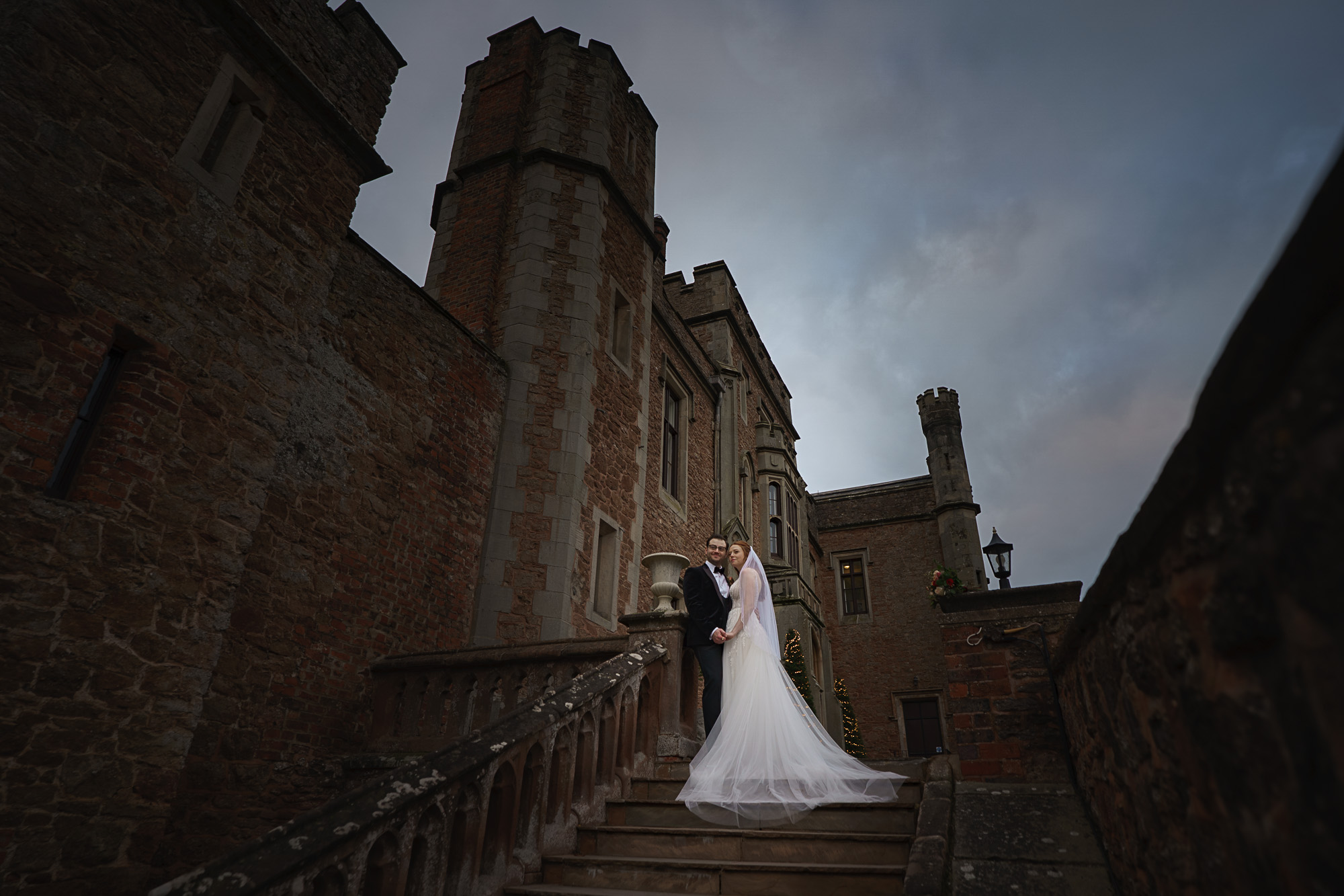 a bride and groom stand together on stone steps outside a large, historic brick building under a cloudy sky, beautifully captured by pbartworks photography, your trusted shropshire wedding photographer.