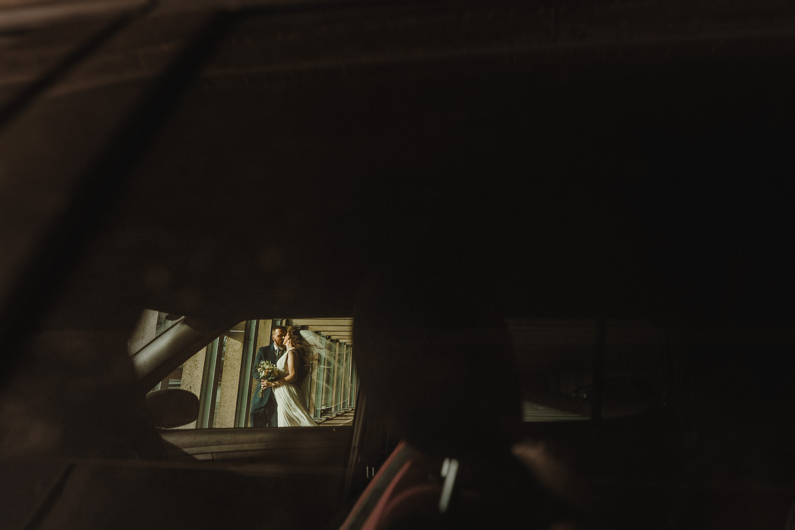 a bride holding a bouquet stands outside, seen through the window of a dark, parked car—captured by a talented shropshire wedding photographer.