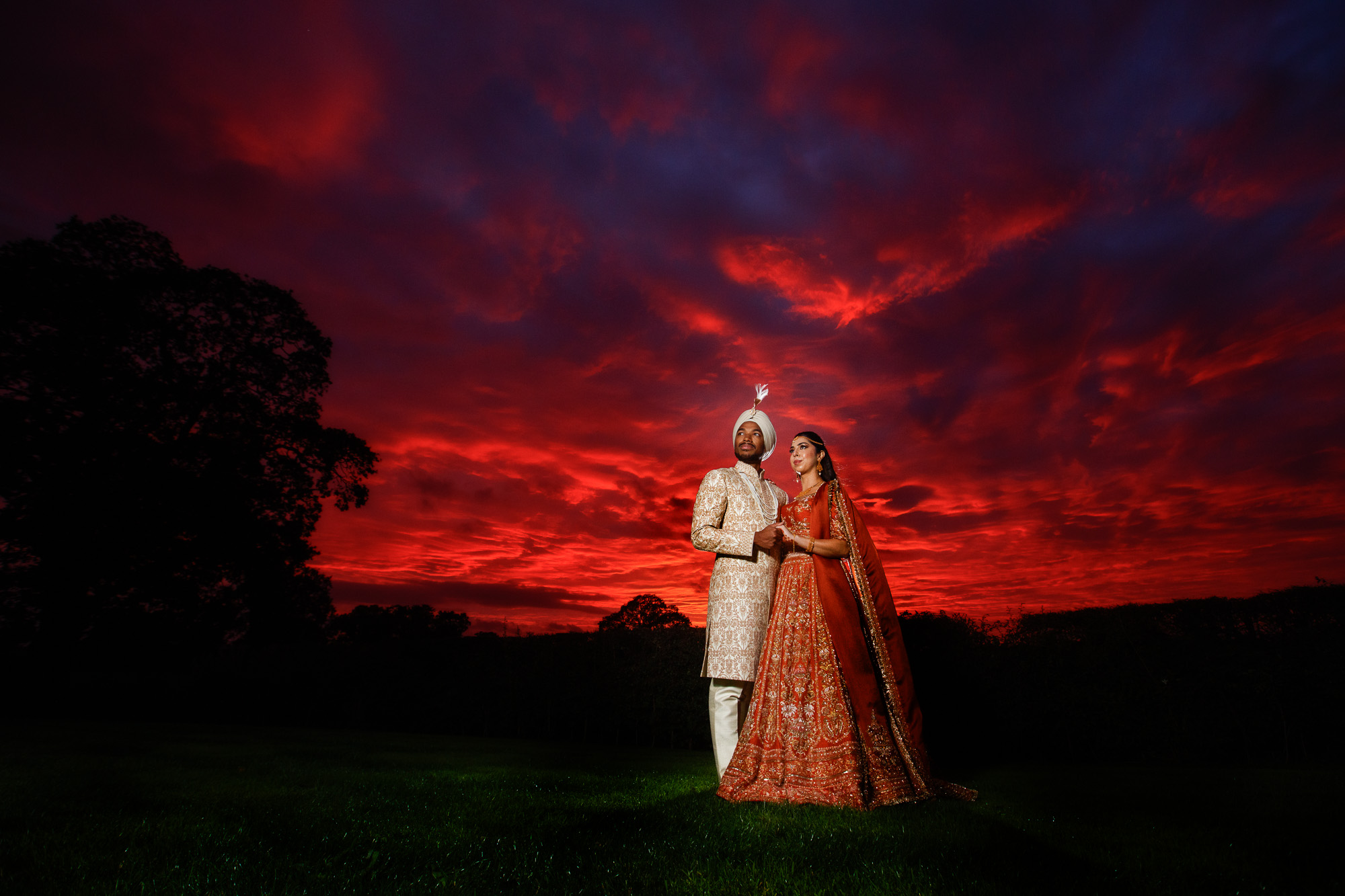 a couple in traditional indian wedding attire stand together on grass at sunset, with a dramatic red and purple sky in the background—perfectly captured by a talented shropshire wedding photographer.